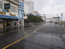 A boardwalk, wet with rain, with shops on the left and a ferris wheel far in the background, the sky overcast